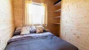 Een kleine kamer met houten wanden, een bed met een geborduurde spreideken, een raam met gordijnen en een wandplank.