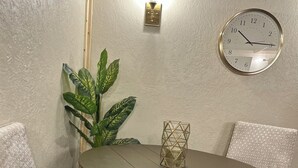 A round table with a light brown top, surrounded by two patterned chairs. A wall-mounted clock is visible above the table.