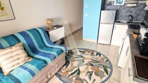 A compact living space with a sofa, a small side table, a kitchen area with a refrigerator, and a patterned rug. - Studio 4 personnes, terrasse vue mer et parking (Sète)