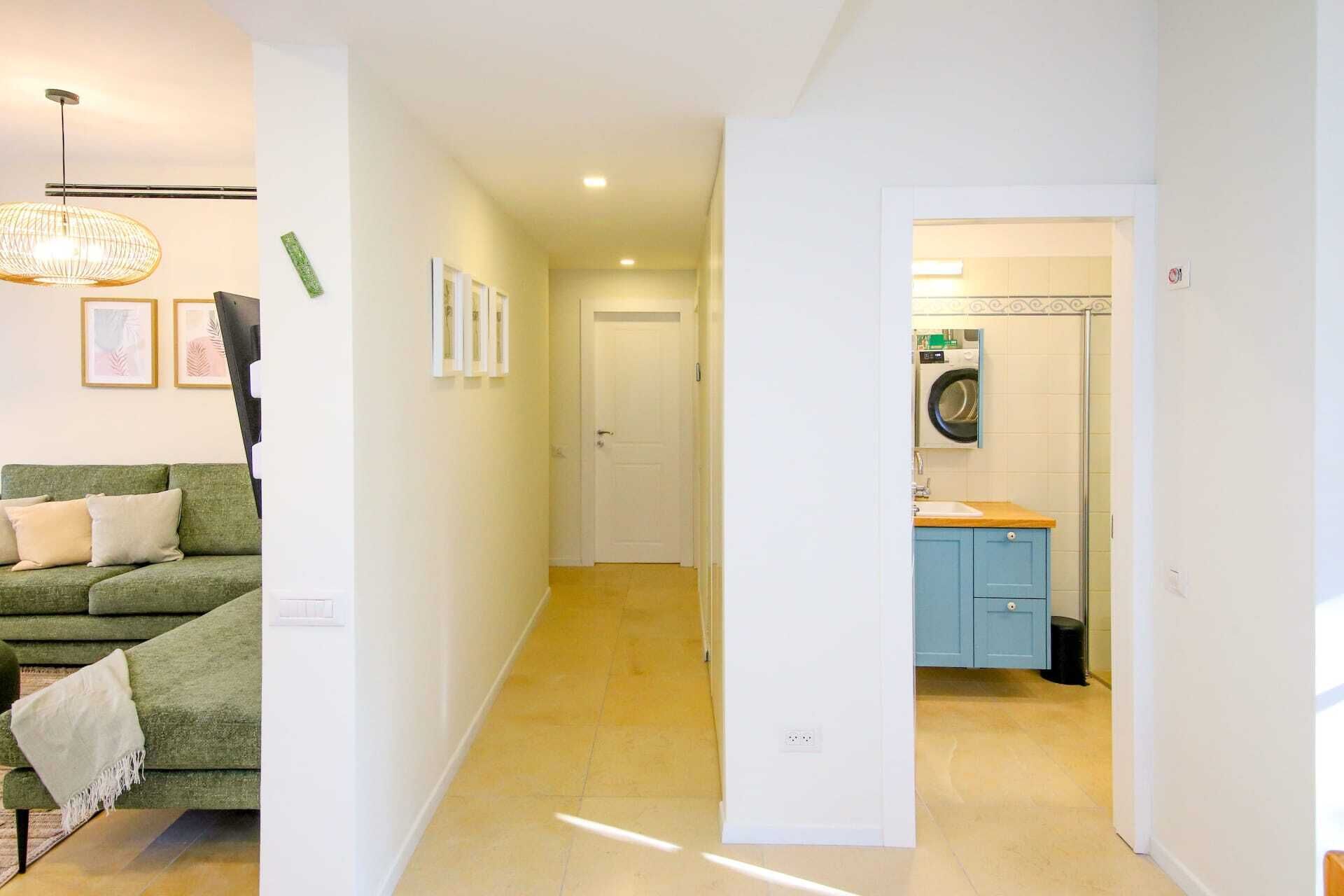 A hallway leading to a bathroom with a washing machine, a green sofa, and framed pictures on the wall.