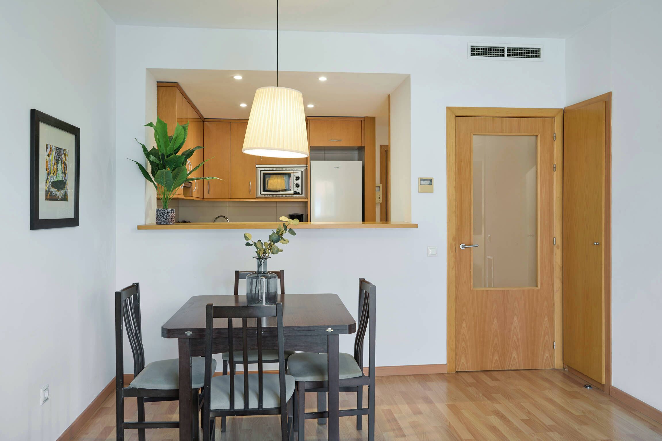 Une cuisine compacte avec des meubles en bois, un four à micro-ondes et un réfrigérateur. On y trouve une table à manger avec des chaises et un luminaire suspendu au-dessus.
