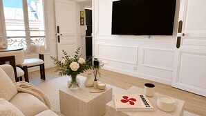A living room with a sofa, a coffee table with a book and candles, a TV on the wall, and a window with a view of buildings.