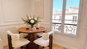 A dining area with a round table, four chairs, a vase with flowers, and a view of buildings through a window.