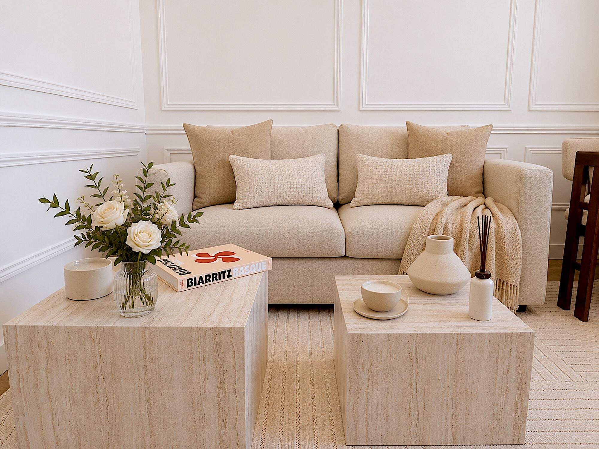 Un salón en tonos neutros con un sofá color beige, dos mesitas auxiliares a juego, un jarrón con flores y un libro titulado 'BIARRITZ BASQUE'.