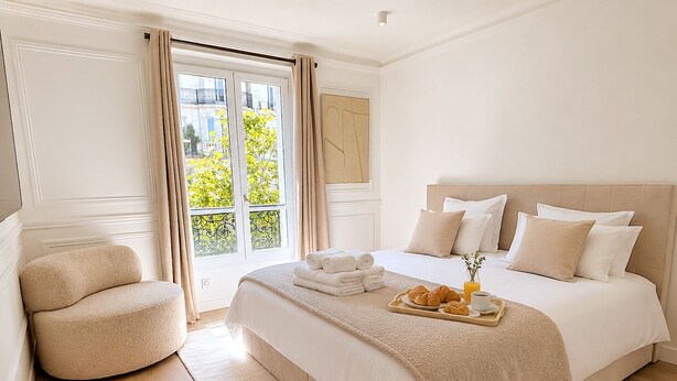 A hotel room with a large bed, a chair, a breakfast tray with croissants, and a view of greenery outside.