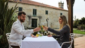 Two people toasting at a table with a white tablecloth, surrounded by greenery and a stone building with a tiled roof in the background. - ABADIA CALDARIA (Arnoia)