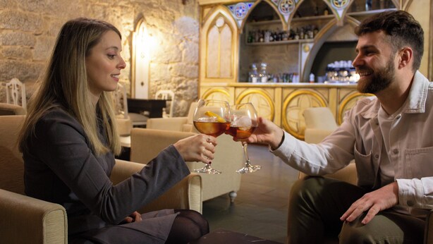 A couple toasting with drinks in a cozy, stone-walled bar area with a bar and shelves of bottles. - ABADIA CALDARIA (Arnoia)