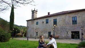 A stone building with a tiled roof, a chimney, and a small tower. There are two windows and a door visible. - ABADIA CALDARIA (Arnoia)