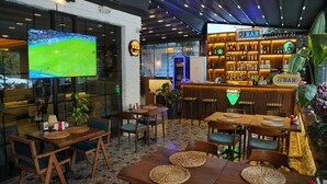 Una zona de bar con mobiliario de madera, un televisor mostrando un partido de fútbol y una variedad de botellas en estanterías.