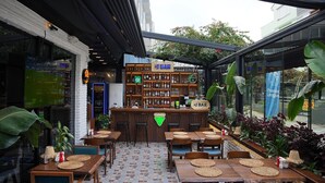 Zona de bar al aire libre con mesas y sillas de madera, barra con un letrero verde y estantes llenos de botellas.