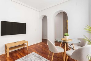 Ein modernes Wohnzimmer mit einem Flachbildfernseher, einer hölzernen Bank, einem Glastisch mit Stühlen und einer kleinen Topfpflanze.