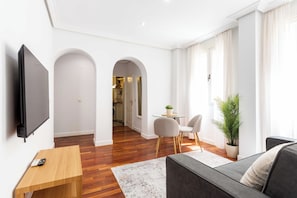 Ein modernes Wohnzimmer mit Flachbildfernseher, Couchtisch aus Holz, grauem Sofa und Essbereich mit zwei Stühlen und einem kleinen Tisch.