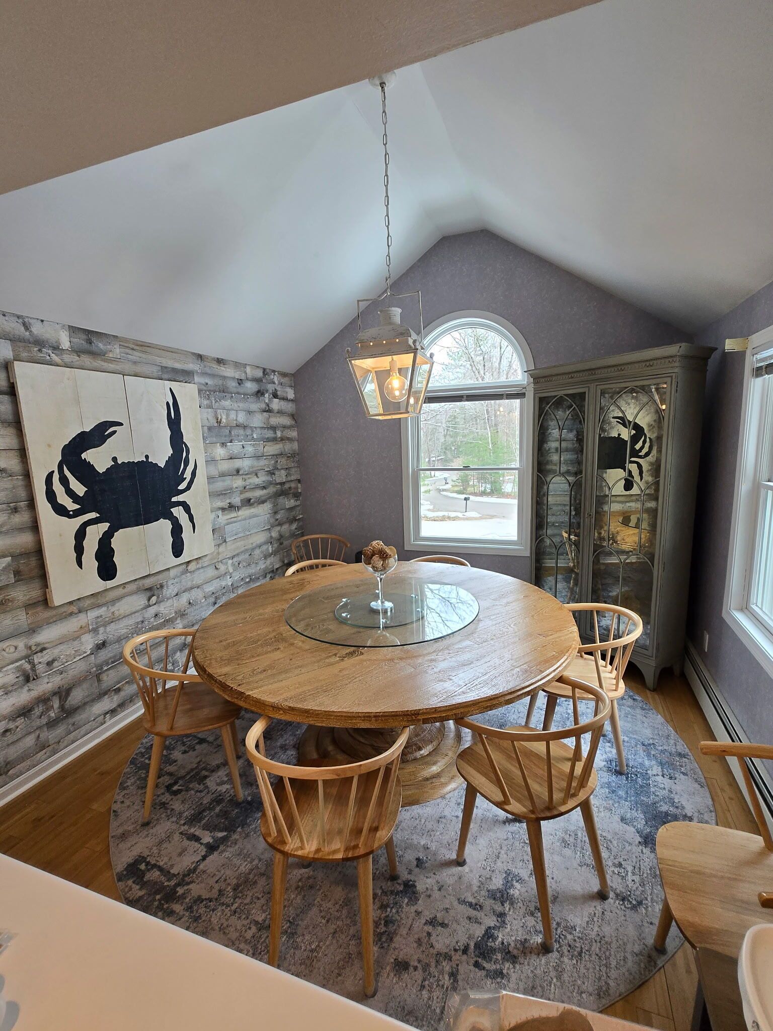 A dining area with a round glass table, wooden chairs, a rustic wall, and a decorative crab painting.
