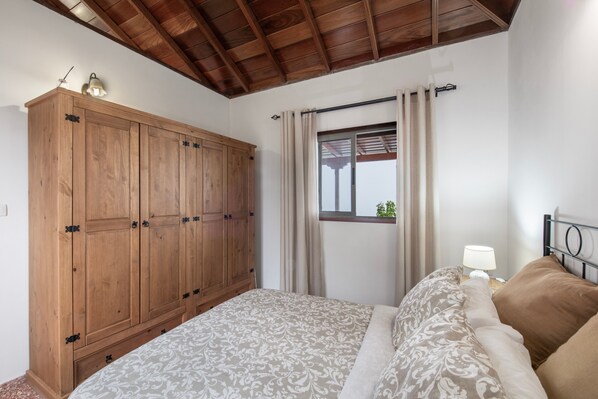 Une chambre avec une armoire en bois, un lit avec une literie à motifs, une fenêtre avec des rideaux et une table de chevet avec une lampe.