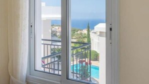 A window with a view of a swimming pool and a building with a white facade. - Akamas Bayview Apartment (Neo Chorio)