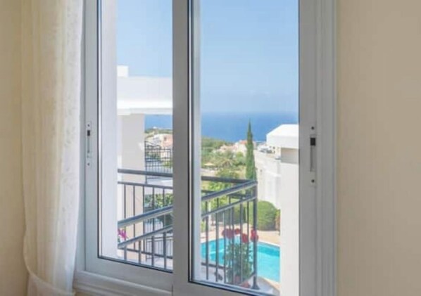 A window with a view of a swimming pool and a building across the water. - Akamas Bayview Apartment (Neo Chorio)