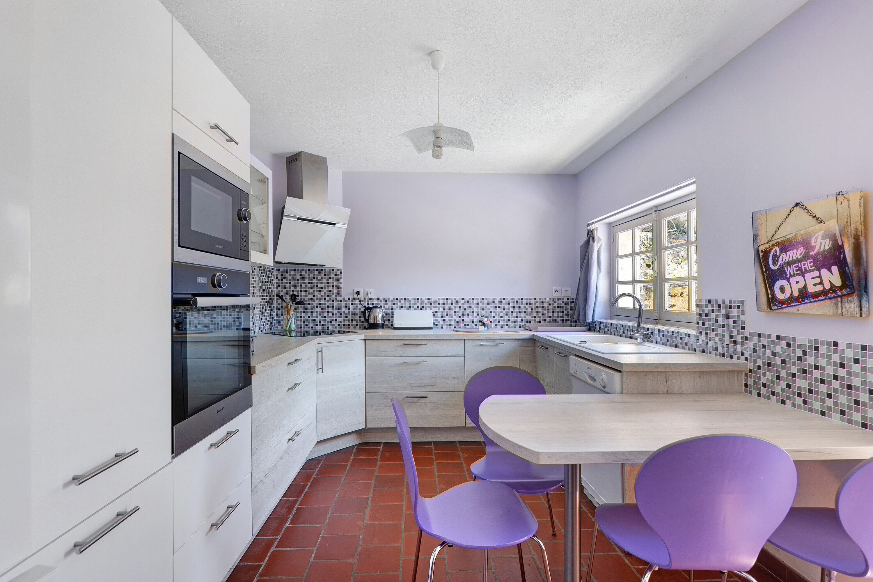 A modern kitchen with white cabinets, a built-in oven, a microwave, and a dining area with purple chairs.