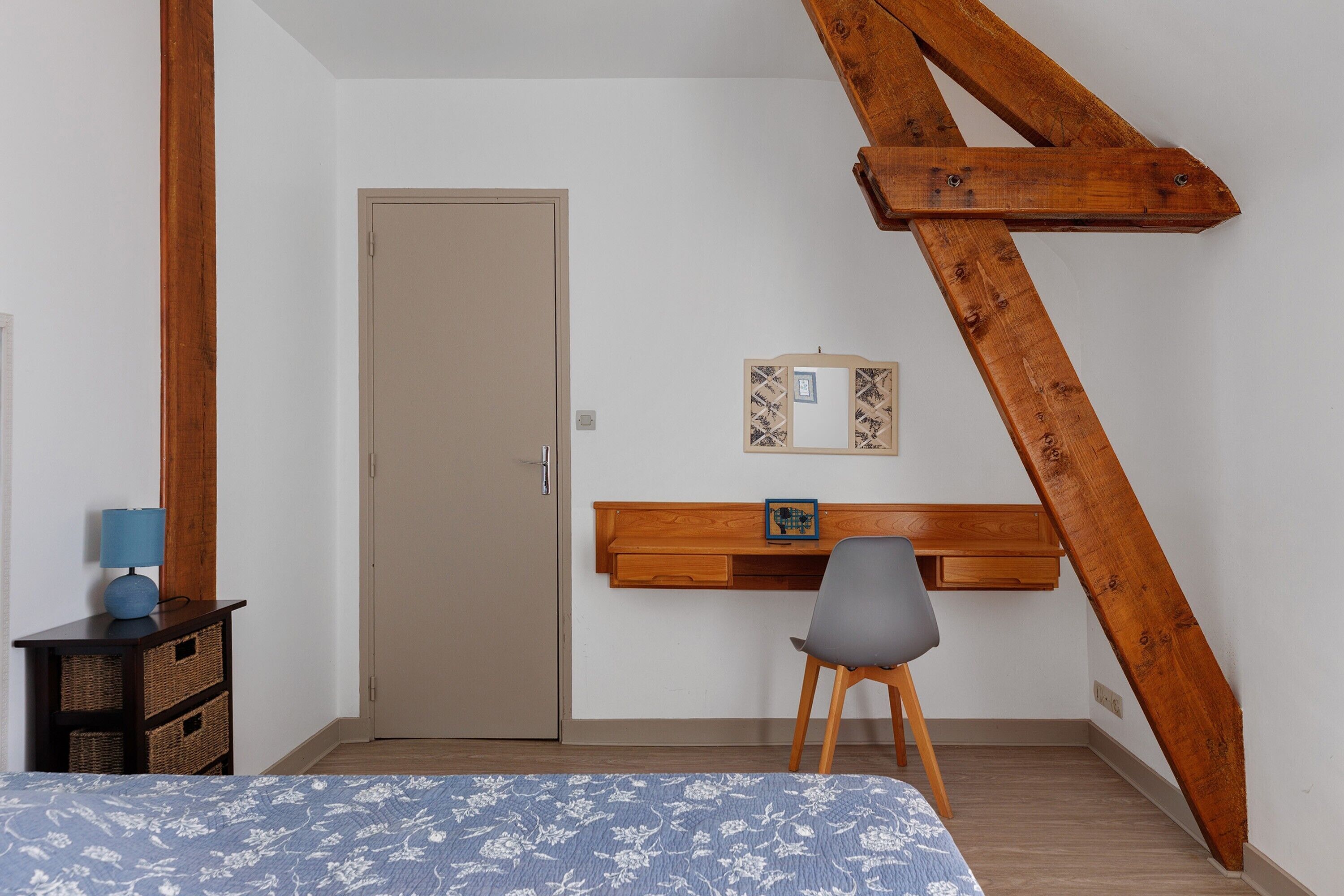 A bedroom with a bed, a desk with a chair, a wooden beam, a door, and a wall-mounted picture.