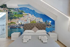 A bedroom with a large bed, two bedside tables, a view of a coastal town through a mural, and a balcony with a railing.