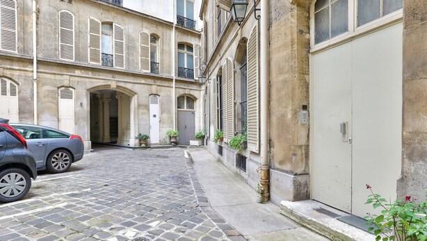 Une rue pavée avec une voiture garée et un immeuble doté de nombreuses fenêtres et volets.
