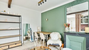 Un salon avec un mur vert, une table à manger en bois, des chaises blanches, un fauteuil en fausse fourrure blanche, une table basse en bois et une bibliothèque.