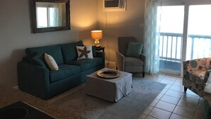 Een woonkamer met een blauwgroene bank, een grijze fauteuil, een kleine tafel met dienblad, een vloerlamp en een raam met gordijnen.