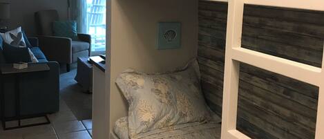 A bunk bed with floral bedding, a chair, and a small table in a room with a window and a view of the outdoors.