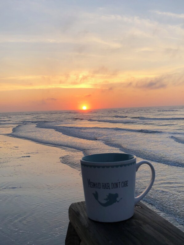 Eine Tasse mit Meerjungfrauen-Motiv auf einer Holzunterlage am Strand bei Sonnenuntergang.