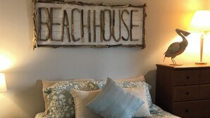 Een slaapkamer met een bed, kussens, een nachtkastje, een dressoir, een lamp en een wanddecoratie met de tekst 'BEACHHOUSE'.