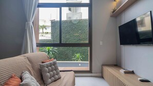 Un salon moderne doté d’une grande fenêtre, d’un canapé avec des coussins, d’un téléviseur à écran plat et d’un banc en bois.