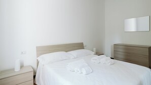 Una cama bien hecha con sábanas blancas y toallas enrolladas, un cabecero de madera, una mesita de noche con una lámpara y un espejo montado en la pared.