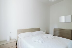 Cama bem arrumada com lençóis brancos e toalhas dobradas, cabeceira de madeira, mesa de cabeceira com luminária e espelho fixado na parede.