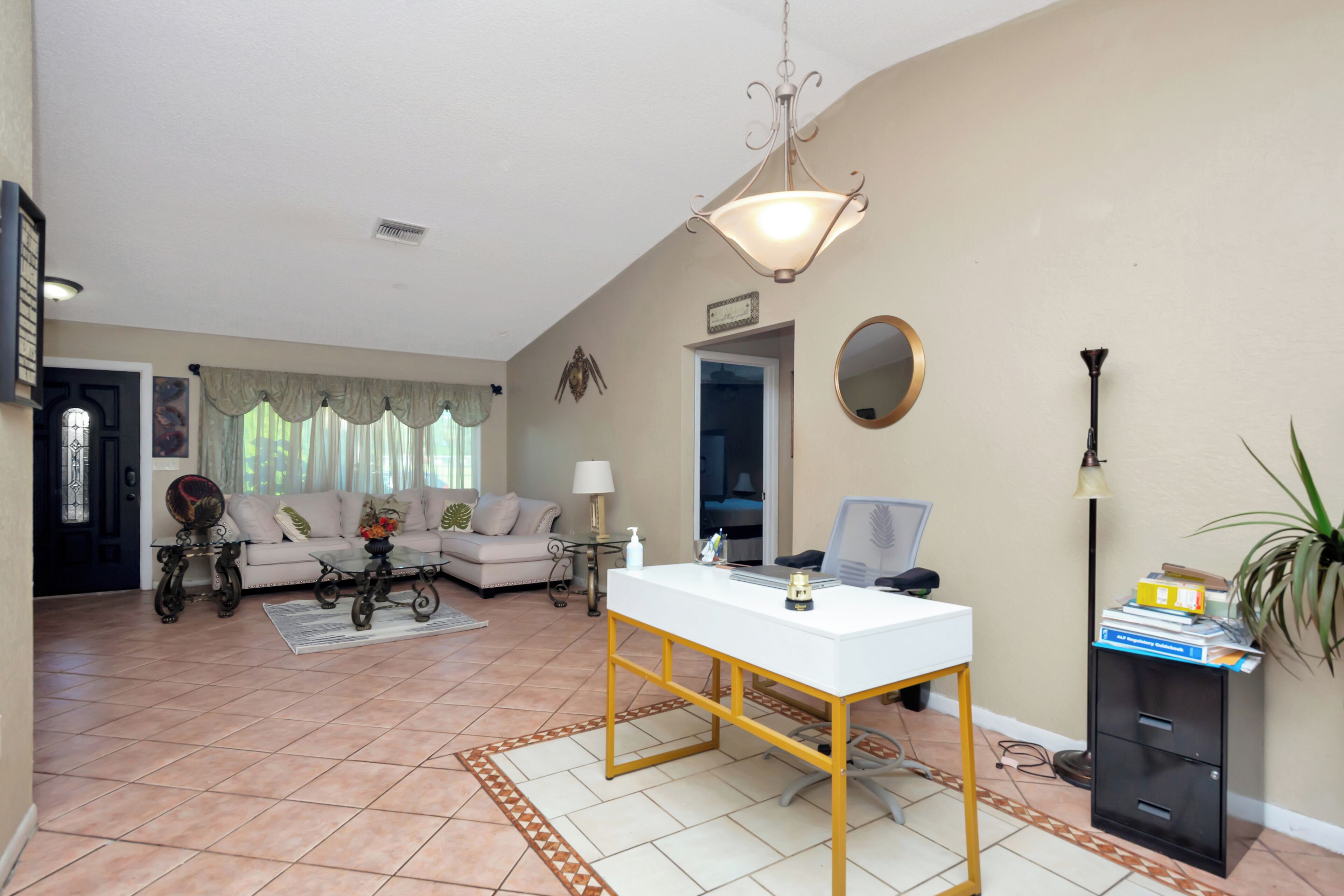 A living room with a white desk, a sofa, a coffee table, and a lamp. There is a door with a decorative glass panel and a plant on the right side.