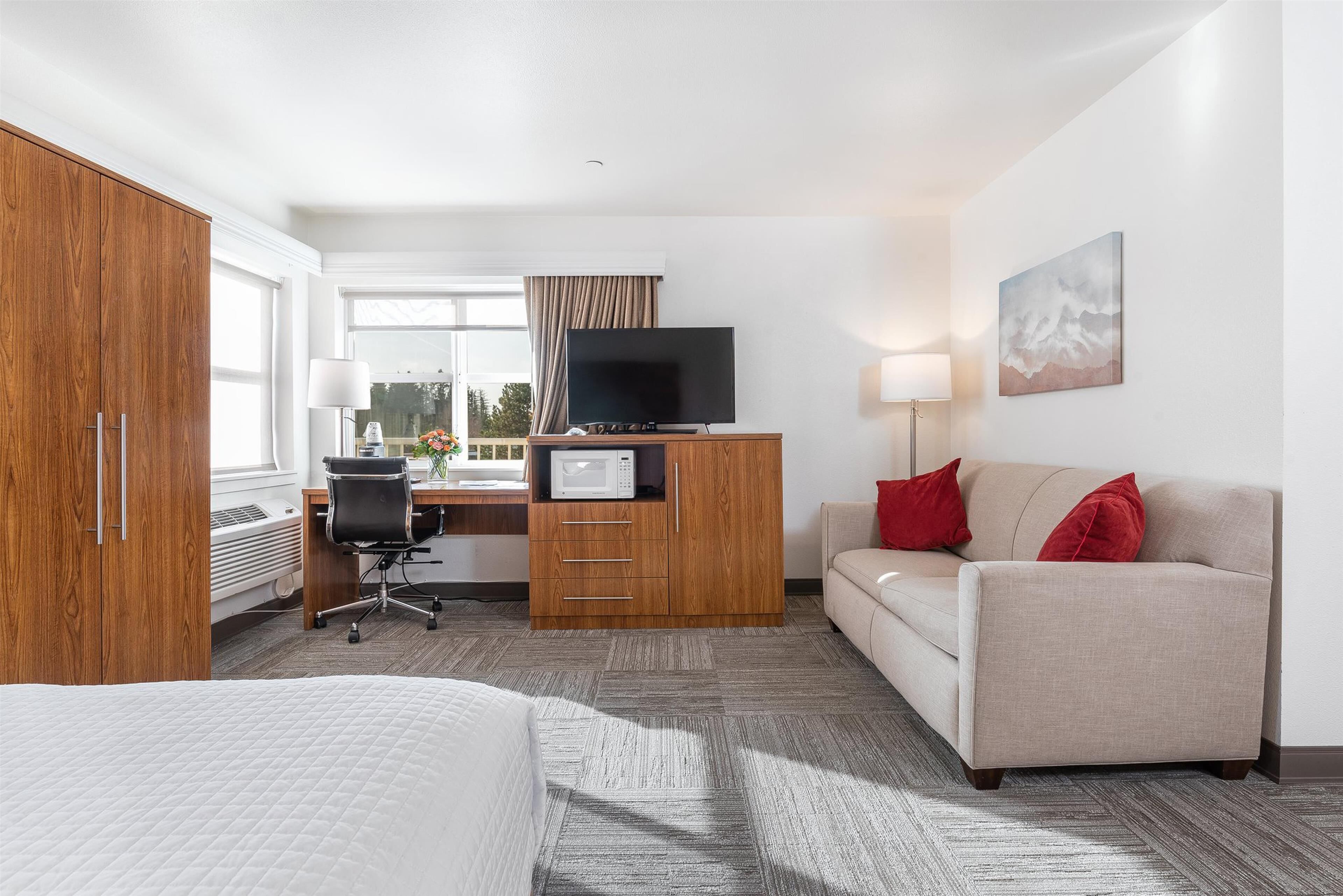 A hotel room with a bed, a desk with a computer, a television, a sofa, and a window with curtains.
