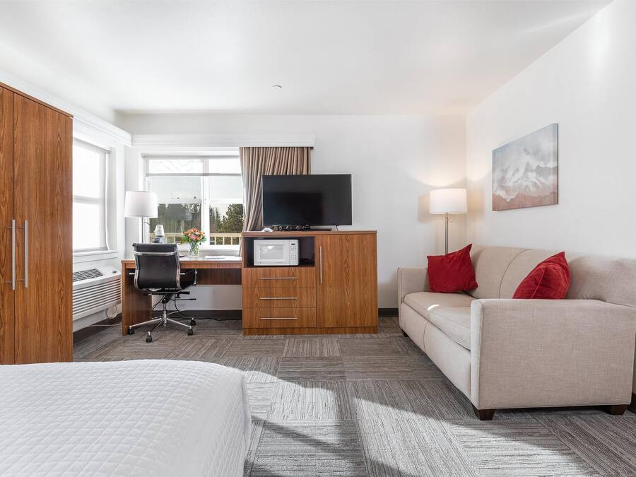 A hotel room with a bed, a desk with a computer, a television, a sofa, and a window with curtains.