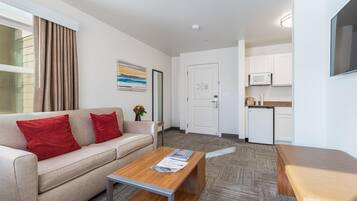 A modern, well-lit room with a sofa, a wooden coffee table, a small kitchen area with a microwave, and a door leading to another room.