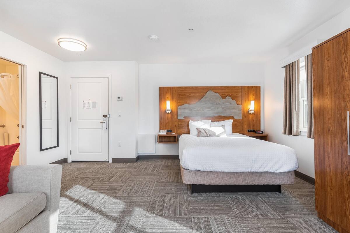 Une chambre d’hôtel avec un lit, une table de chevet, une tête de lit en bois, une lampe fixée au mur et une porte.