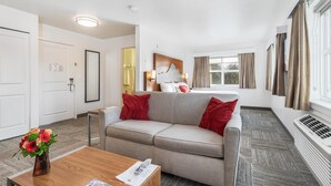 Une chambre d’hôtel moderne avec un canapé gris, des coussins rouges, une table basse en bois et une grande fenêtre avec des rideaux.