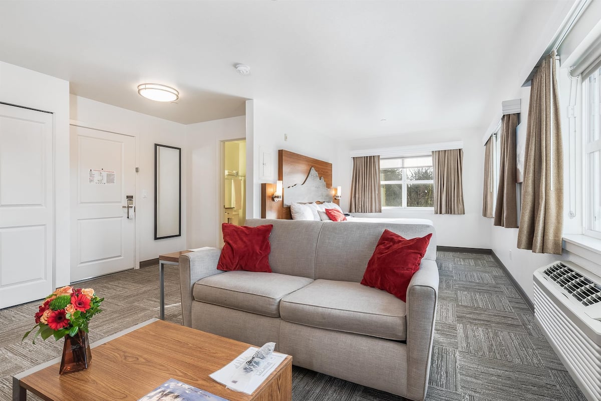 Une chambre d’hôtel moderne avec un canapé gris, des coussins rouges, une table basse en bois et une grande fenêtre avec des rideaux.