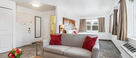 Ein modernes Hotelzimmer mit grauem Sofa, roten Kissen, einem Holztisch und einem großen Fenster mit Vorhängen.