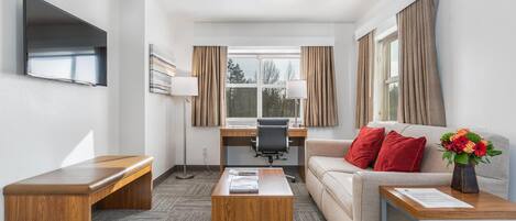Una habitación de hotel moderna con sofá, una mesita de centro de madera, un escritorio con silla, televisor de pantalla plana montado en la pared y una ventana con cortinas.