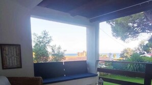 Quarto com vista para um jardim e uma varanda. Há um sofá de vime, uma mesa de centro de madeira com uma fruteira e uma almofada branca.