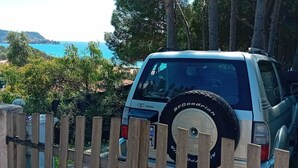 Carro estacionado numa área cercada, com vista para o mar e árvores.