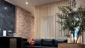 A modern living room with a black leather sofa, a wooden coffee table, and a white rug. There are three orange suitcases stacked in the corner.