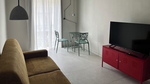A living room with a sofa, a dining table with chairs, a TV on a red cabinet, and a large window with sheer curtains. - Fermata Marconi two-room apartment in the center of Turin (Torino)