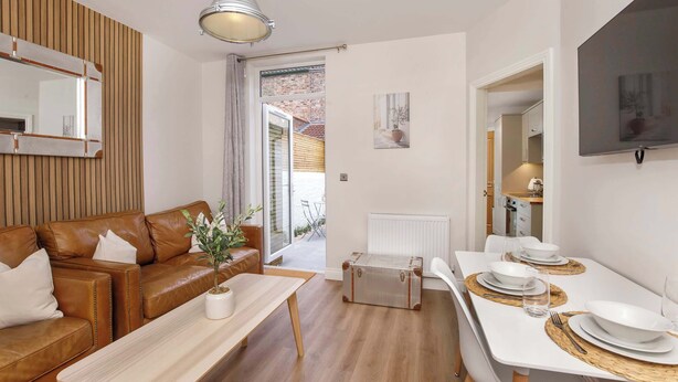 A modern living room with a brown leather sofa, a wooden coffee table, and a dining area with a white table set for two. - The Granville, 4 bedroom house, sleeps 7 (York)
