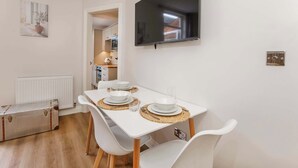 A dining area with a white table set for four, a television mounted on the wall, and a suitcase on the floor. - The Granville, 4 bedroom house, sleeps 7 (York)