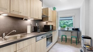 Une cuisine moderne avec des armoires en bois, un évier en acier inoxydable, un four à micro-ondes et un four encastré. On y trouve également deux tabourets verts et une poubelle.