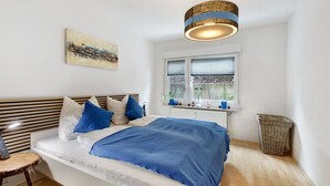 Une chambre à coucher comprenant un lit, une table de chevet, une lampe, une fenêtre avec des stores, une œuvre d’art accrochée au mur et un luminaire suspendu.