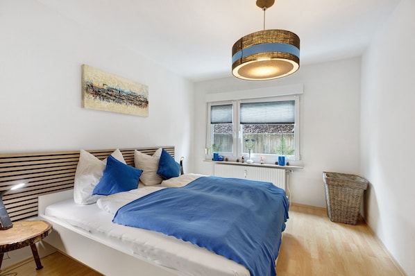 Un dormitorio con cama, mesita de noche, lámpara, ventana con persianas, una obra de arte en la pared y una lámpara colgante.